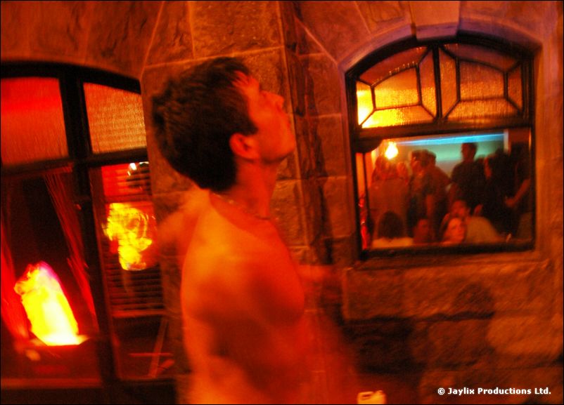 A shirtless man looks up beside a stone window with flames and people visible through the glass