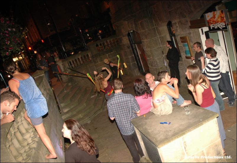 People gather on a night-time terrace by a stone ledge, with a FIRE sign in the background