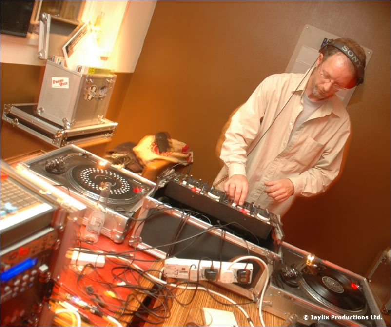 A man in a beige shirt and headphones stands at turntables and a mixer, with flight cases behind
