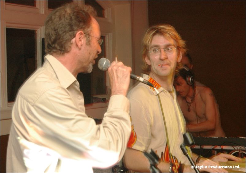 A man in a beige shirt sings into a microphone beside a man with glasses playing a keyboard