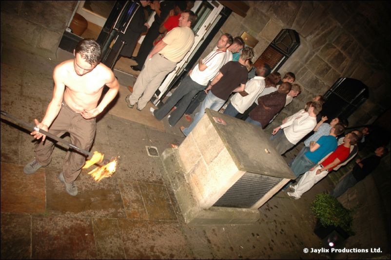 A shirtless man holds a fire staff on a street at night as onlookers stand by a stone wall