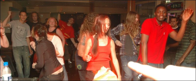 People dance in a room, a man in a red t-shirt with his arm raised at right