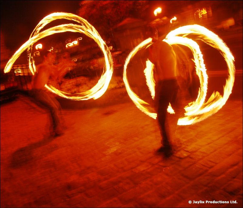 Two people spin rings of fire on a cobbled area at night