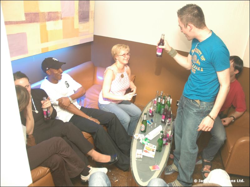 A man in a blue t-shirt raises a bottle beside a table of bottles, with people seated on sofas