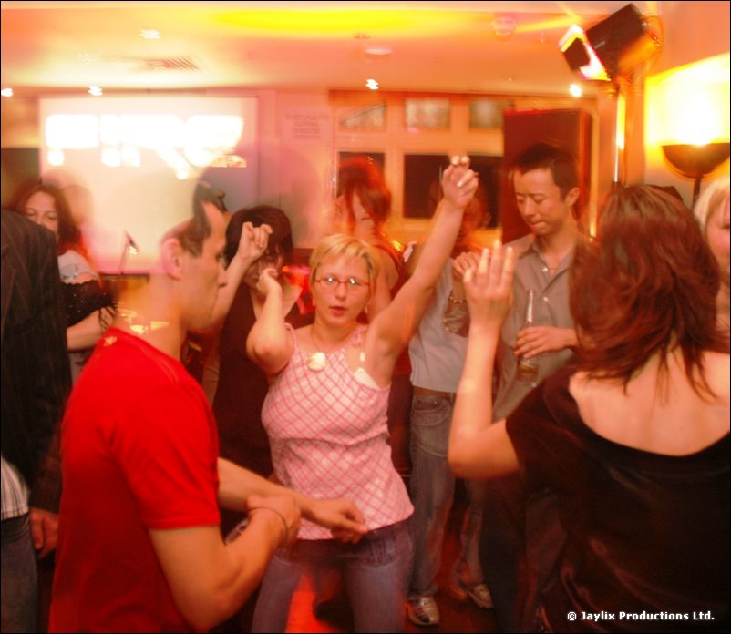 People dancing in a busy, warmly lit bar, a woman in a pink top raising one arm