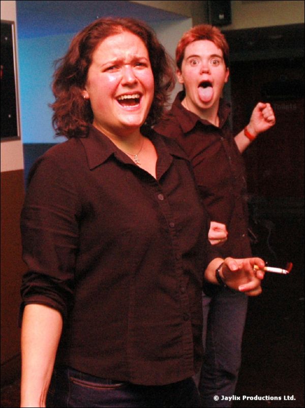 Two people in dark shirts pull expressive faces, one sticking out a tongue, one holding a cigarette