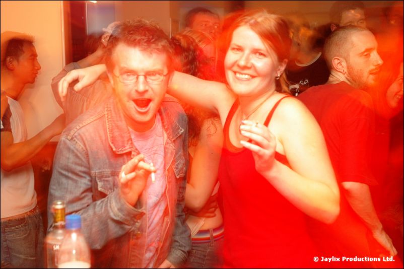 A man with glasses and a woman in a red top dance in a crowd, blurred in red light