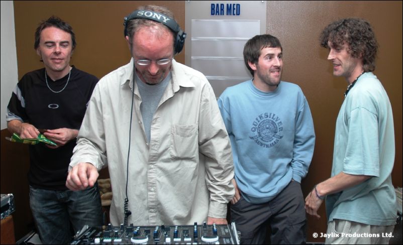 Four men stand around a DJ in headphones at decks, a Bar Med sign on the wall behind