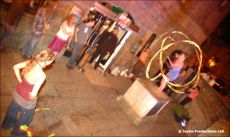 A crowd stands in a stone-paved street at night as a person spins a ring of fire