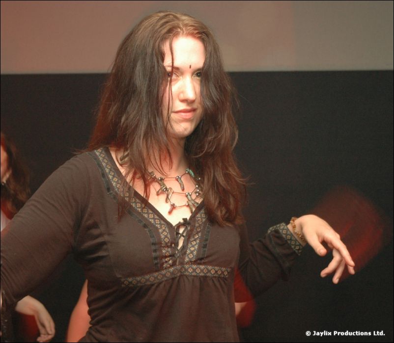 A woman with long dark hair looks at the camera, one hand raised, against a dark wall
