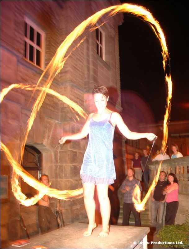 A woman in a blue dress stands on a table tracing rings of fire, people watching beside a stone building
