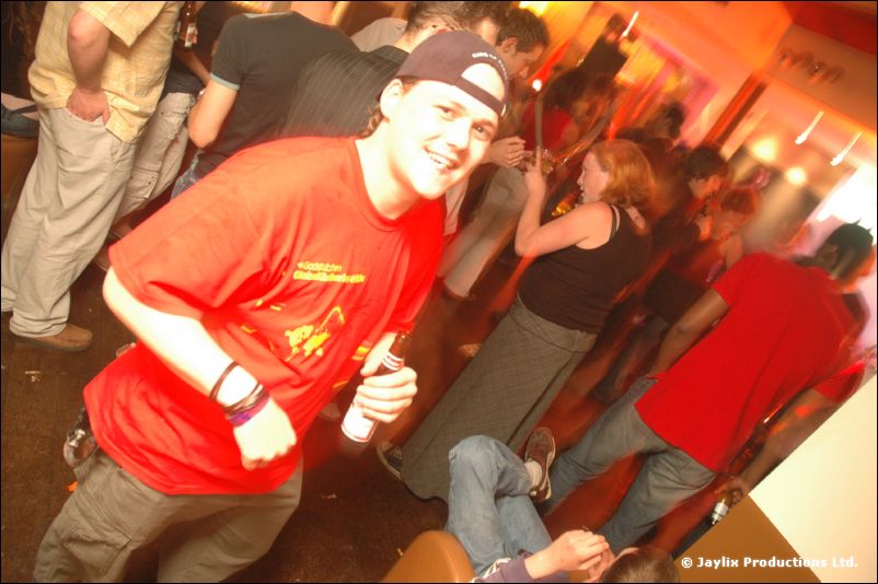 A smiling person in a red t-shirt and cap leans towards the camera in a busy bar