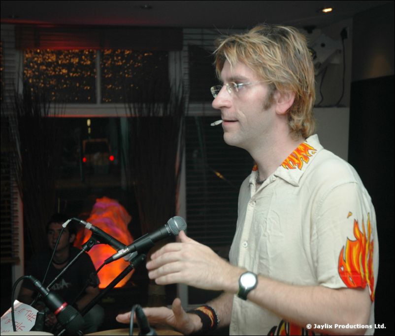 A man with glasses and a cigarette in his mouth talks into a microphone beside drums