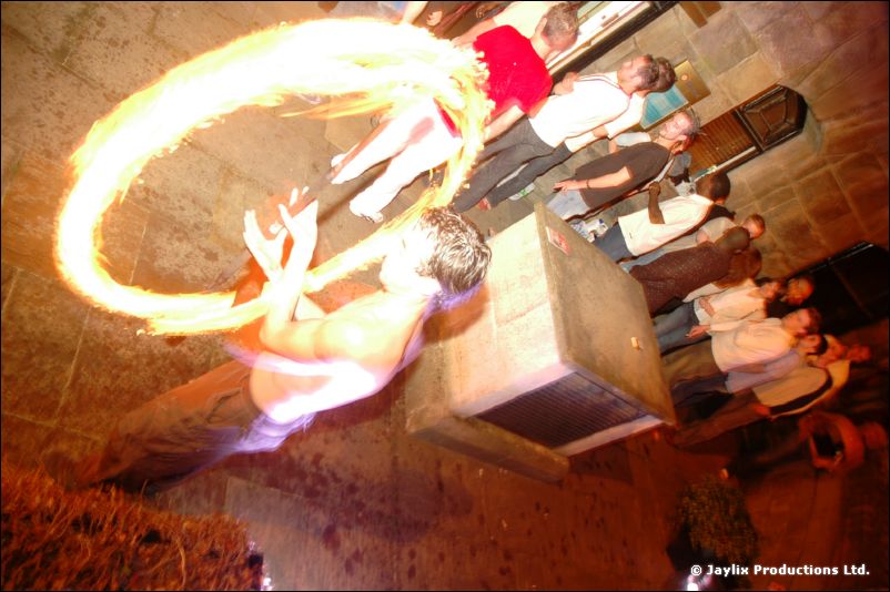 A person with a bare chest spins rings of fire in a street, people standing behind