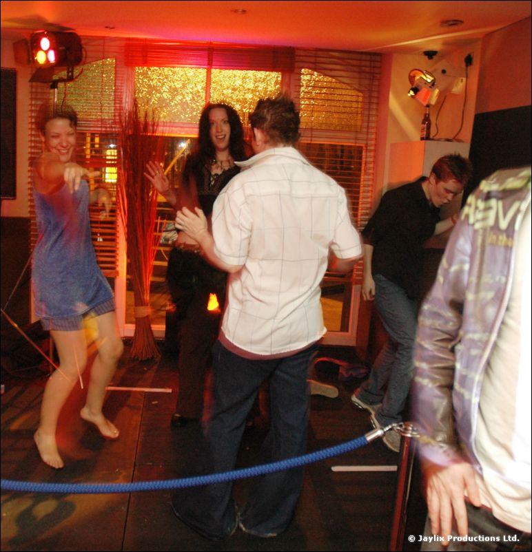 A barefoot woman in a blue dress dances in a bar, people standing around a blue rope