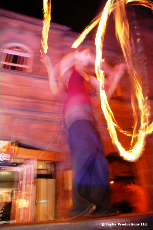 A person spins rings of fire on a table in front of a street of old buildings