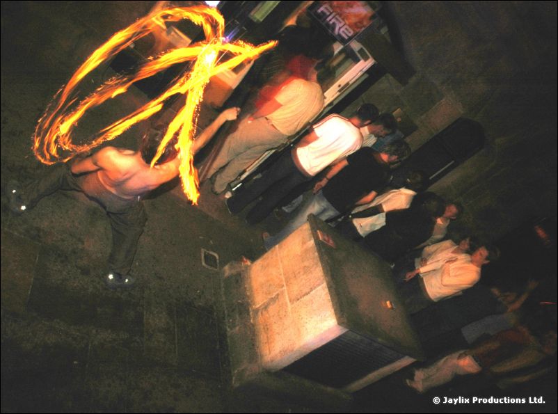 A person spins rings of fire on a stone pavement, people sitting on steps behind