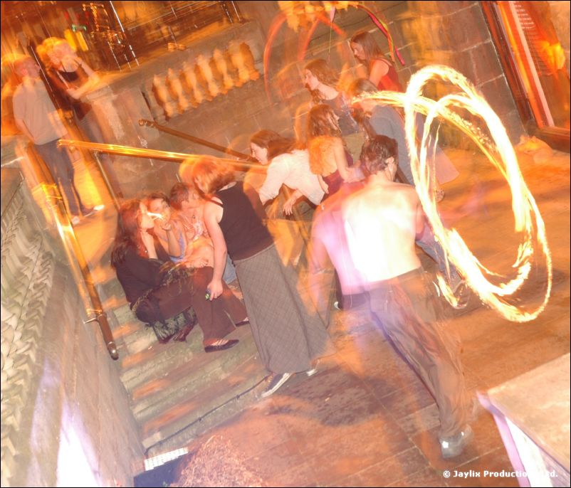 A person spins rings of fire on stone steps beside a crowd sitting on a bench