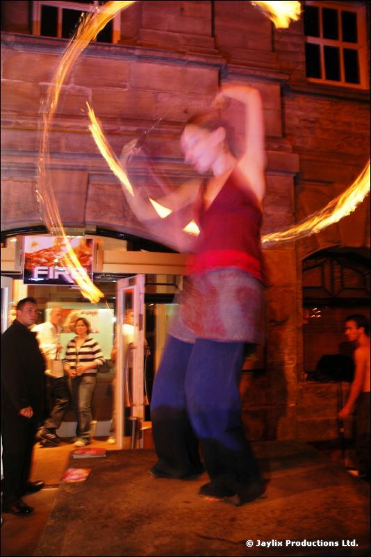 A woman in a blue skirt spins rings of fire in front of a FIRE sign at a doorway