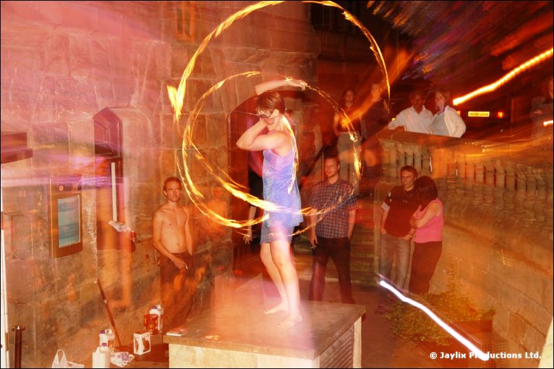 A person in a blue dress spins rings of fire on a table, onlookers around her