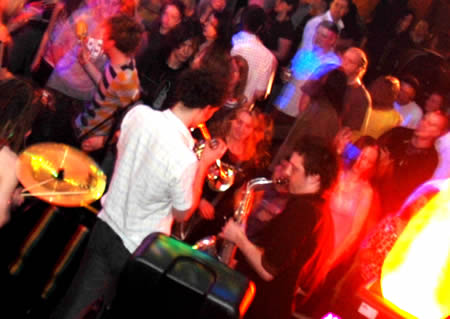 Brass players and a drummer perform among a packed, colourfully lit crowd