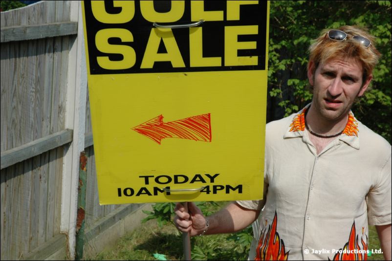 A person in a patterned shirt holds a yellow Golf Sale sign in a garden