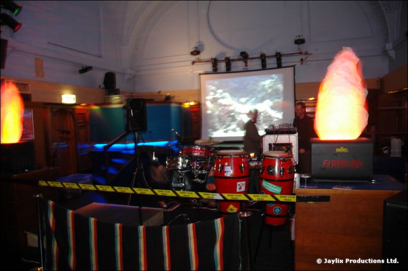 A hall with a projection screen, red conga drums and two glowing flame-effect lights, yellow warning tape strung across the front
