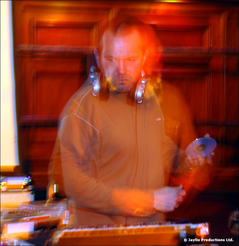 A blurred DJ with headphones holds a CD above decks, lit in orange