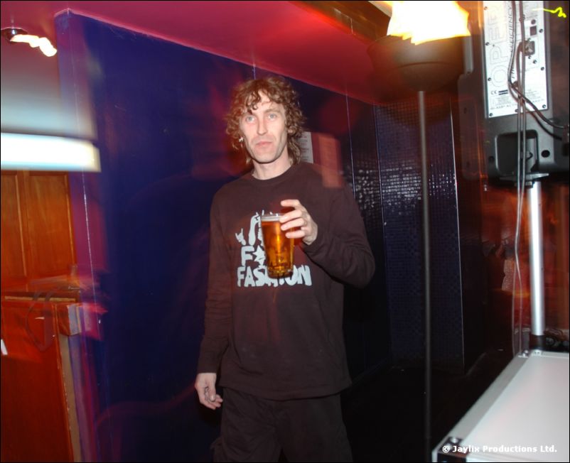 A man with curly hair in a dark T-shirt holds a pint glass in a dim, colourful corner