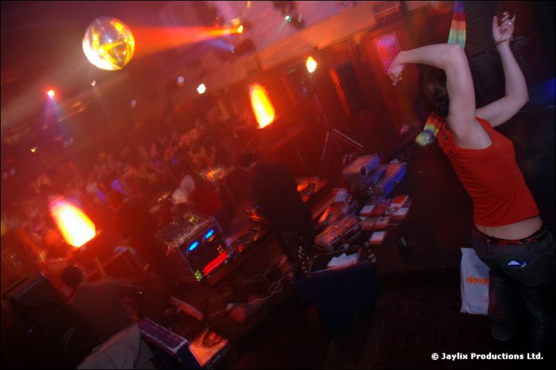 A dancer in a red top raises their arms near DJ decks under a mirror ball