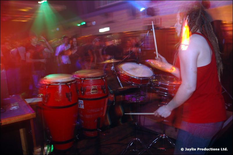 A drummer in a red top plays congas and a drum kit on a busy stage