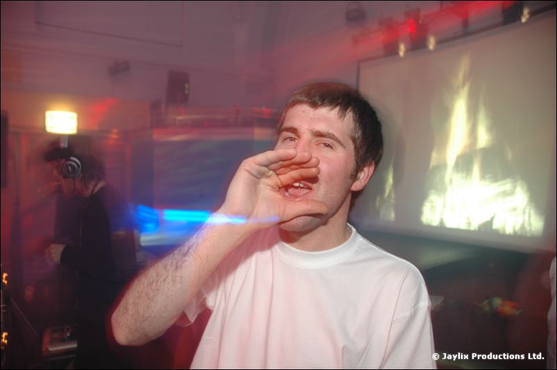 A man in a white T-shirt cups his hands to shout, with a screen showing flames behind