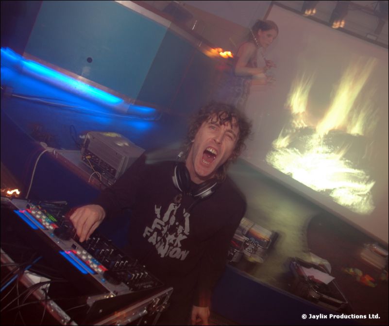 A DJ with headphones shouts at the camera behind mixing equipment, with flames on a screen