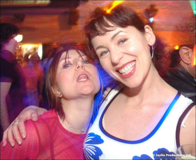 Two women smile and lean together in a busy club, one in a white and blue top