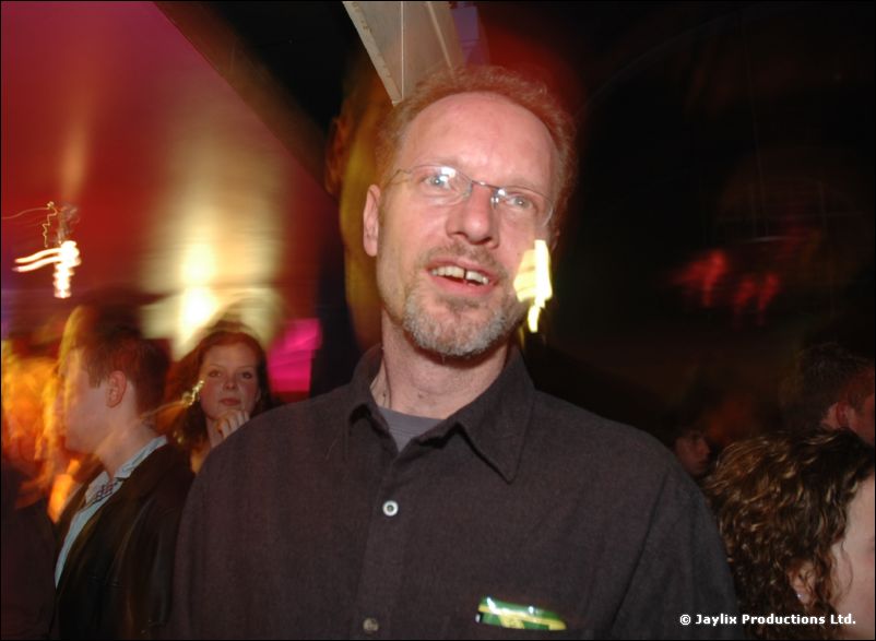 A bearded man with glasses in a dark shirt looks at the camera in a dimly lit club