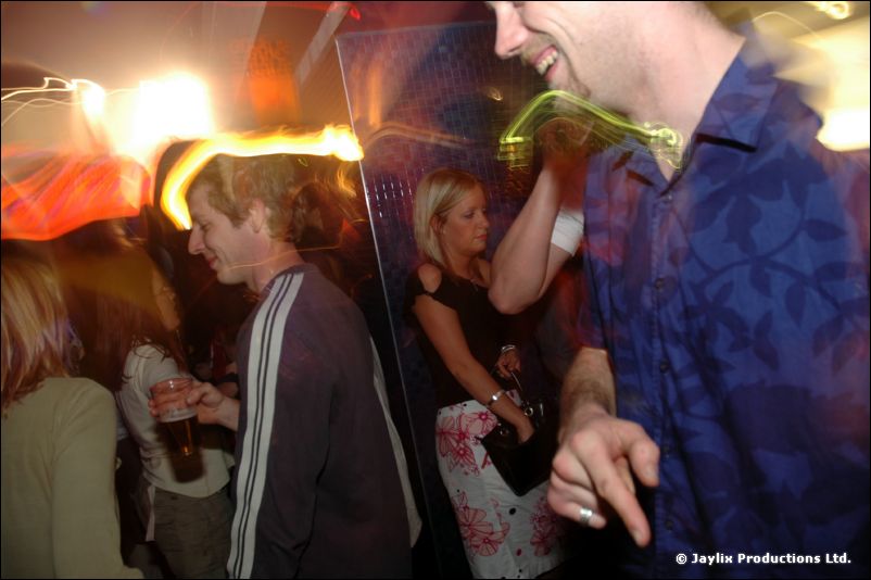 Two men dance in a crowded club with blurred light trails, a woman standing behind