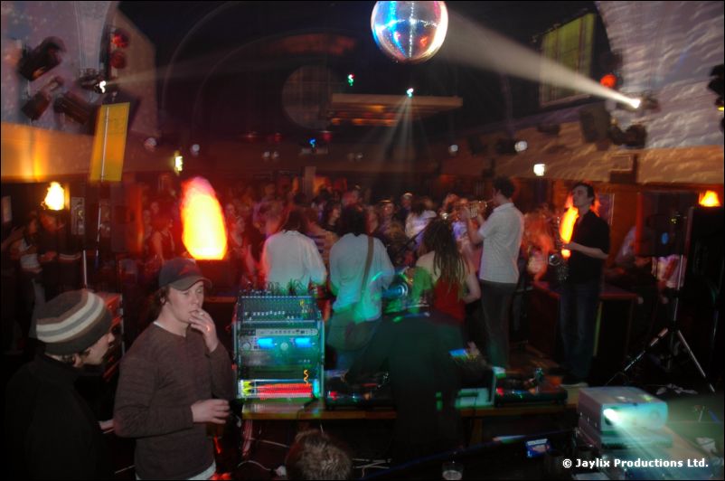 A DJ area with a mirror ball and flame effect, and people standing around in a large room