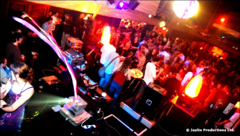 Wide-angle view of a packed club with a band, DJ decks and a glowing flame unit
