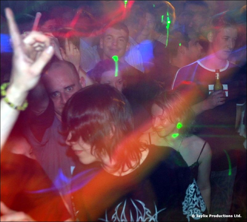 Dancers in a crowd with raised hands under green laser light, one in a black patterned T-shirt