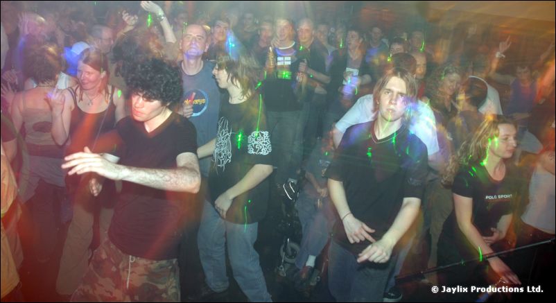 A crowd dances under coloured lights, with a curly-haired person in a dark T-shirt in front