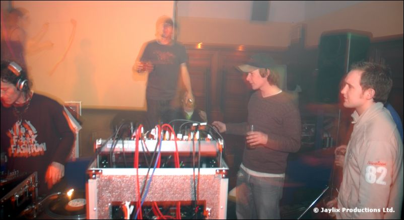 DJs work at a table of mixing gear with people standing around a lit room