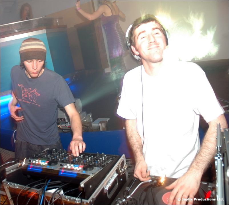 Two DJs at mixing decks, one in a striped beanie and one in a white T-shirt and headphones, smiling
