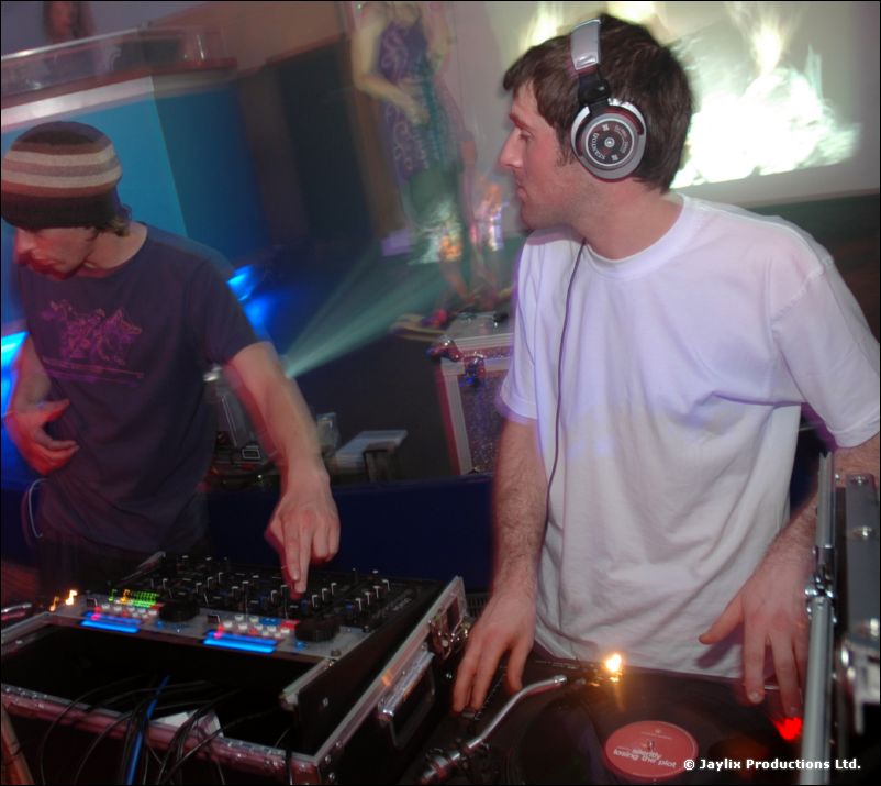 Two DJs at decks, one in a beanie and one in a white T-shirt and large headphones