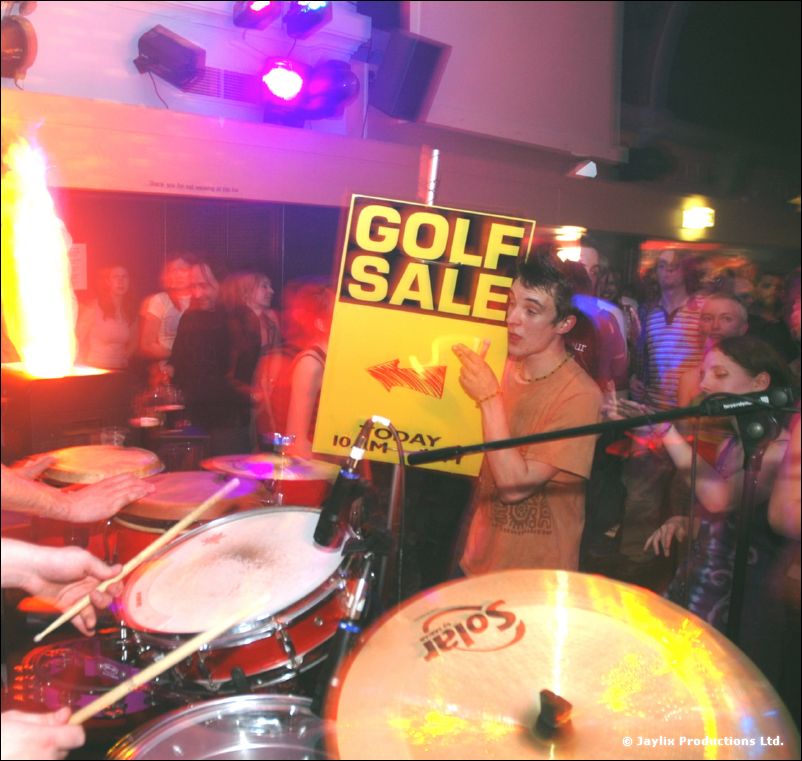 A person in an orange top holds a Golf Sale sign behind a drum kit, with a crowd beyond