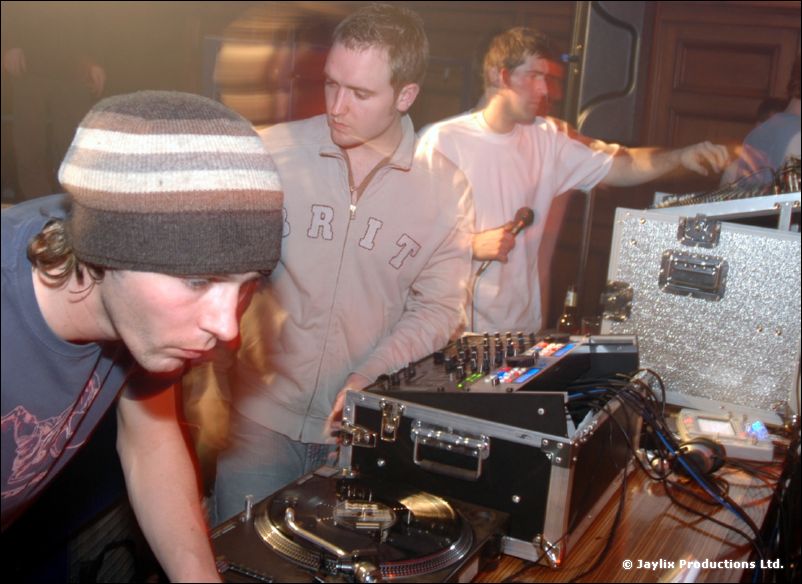 Three DJs at turntables and a mixer, one wearing a beanie and another holding a microphone