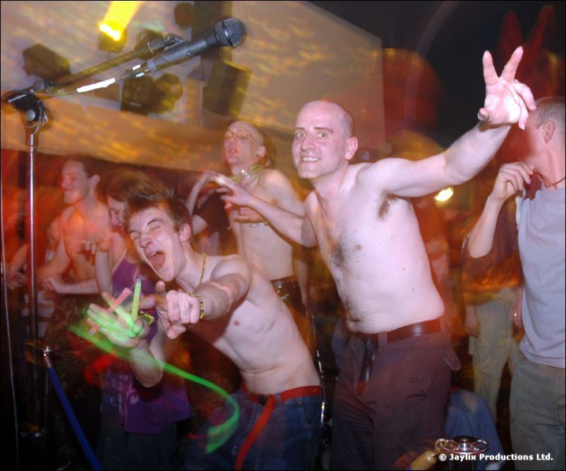 Shirtless men cheer and gesture at the camera in front of a microphone, one holding a glow stick