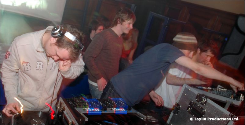 DJs bend over mixing decks, one in a grey hooded top with headphones, another in a beanie