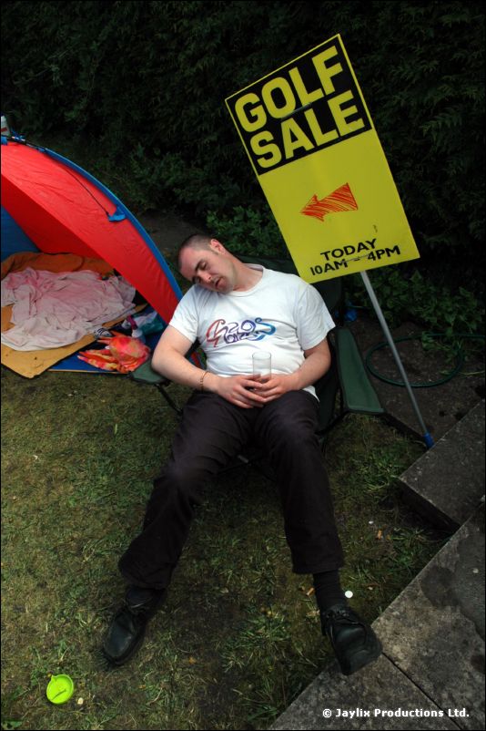 A man dozes in a folding chair beside a small tent and a yellow Golf Sale sign