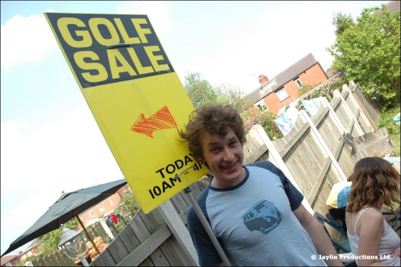 A person in a blue T-shirt holds a Golf Sale sign in a pub garden, with someone behind