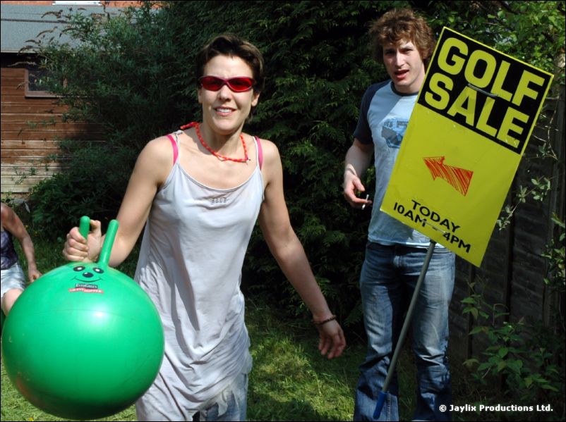 A woman in red sunglasses holds a green space hopper in a garden, with a man holding a Golf Sale sign